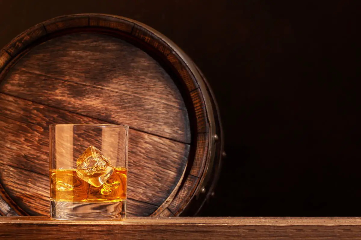 Difference Between Scotch and Bourbon - Scotch on the rocks in front of an oak barrel. 